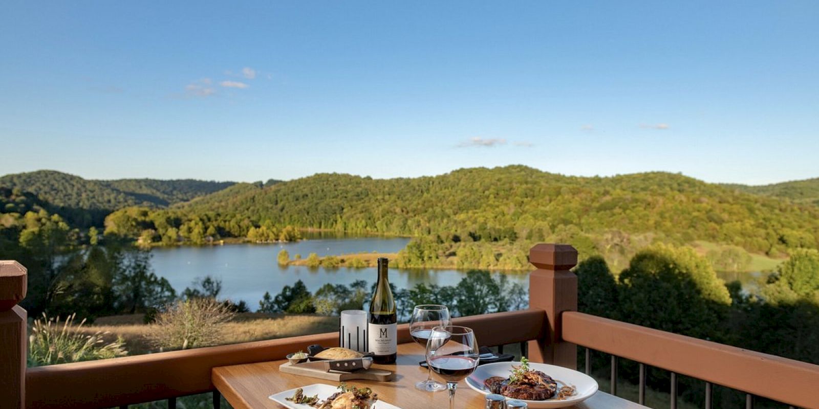 A wooden table with dishes, wine, and glasses set up for dining, overlooking a scenic view of a lake and green hills under a clear blue sky.
