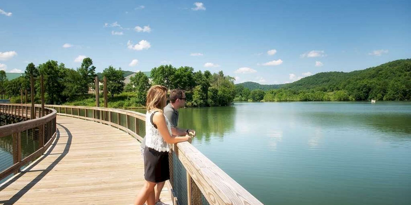 A couple stands on a wooden boardwalk overlooking a serene lake, surrounded by green hills and a clear blue sky with a few white clouds.