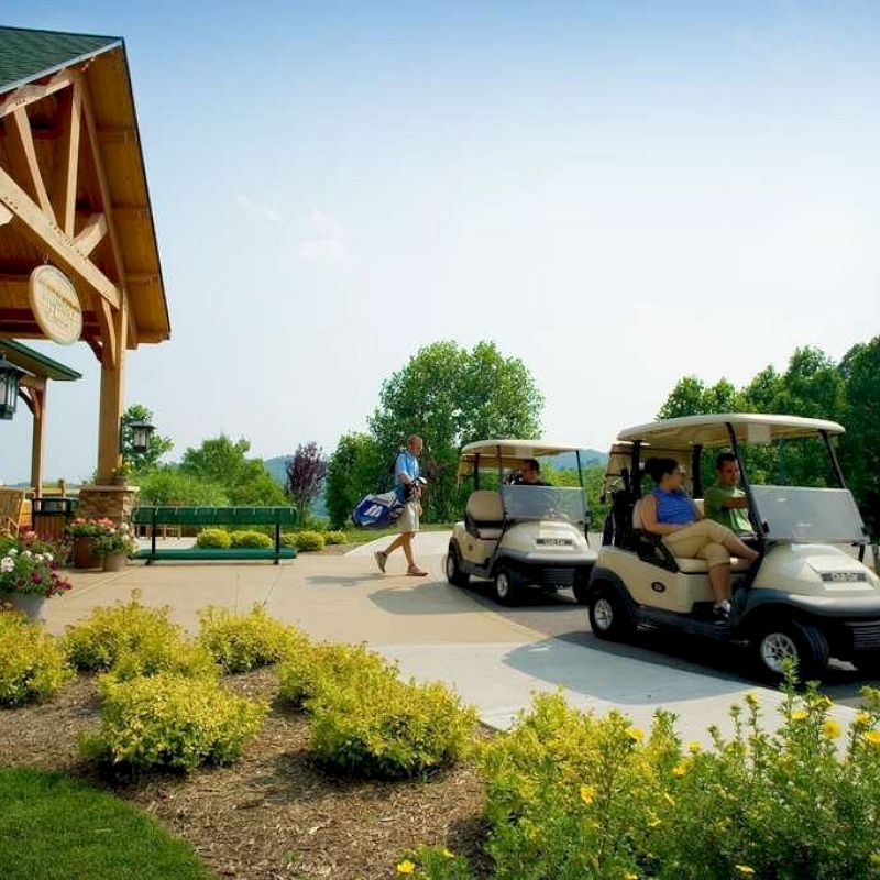 People in golf carts near a building with yellow flowers in front, and a person carrying a golf bag. Trees and clear sky are in the background.