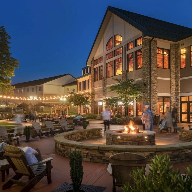 An outdoor area at night with people enjoying a fire pit surrounded by seating, string lights, and a multi-story building with large windows.