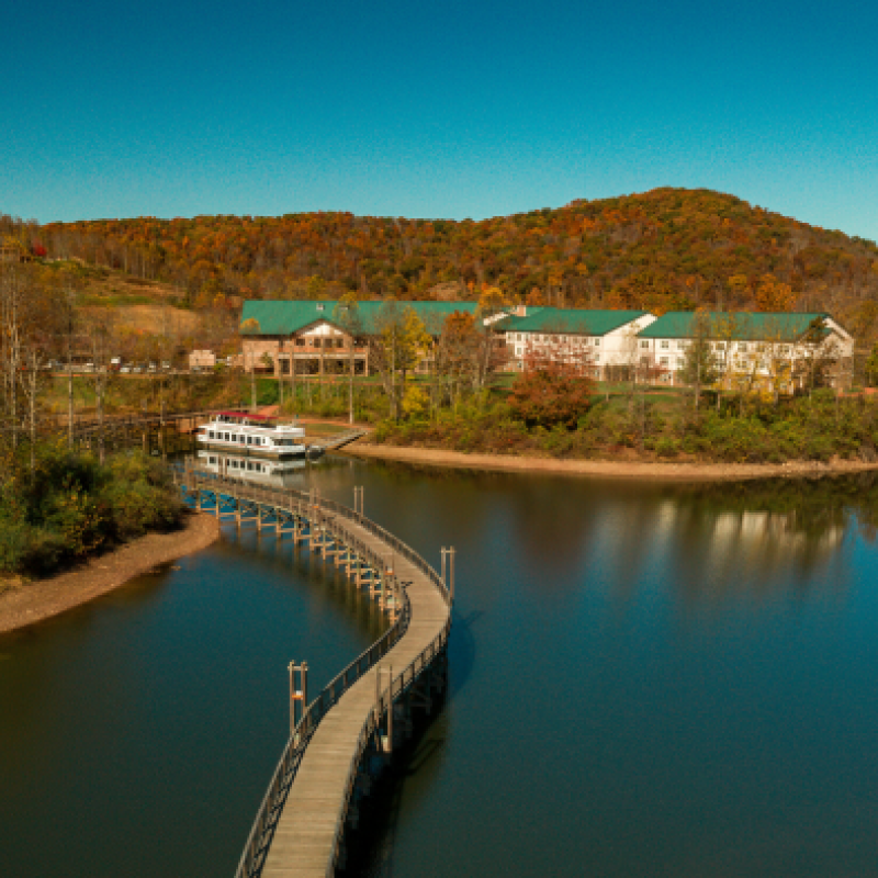 The image shows a scenic lake with a wooden boardwalk leading to buildings on a small island, surrounded by autumn foliage and rolling hills under a clear blue sky.