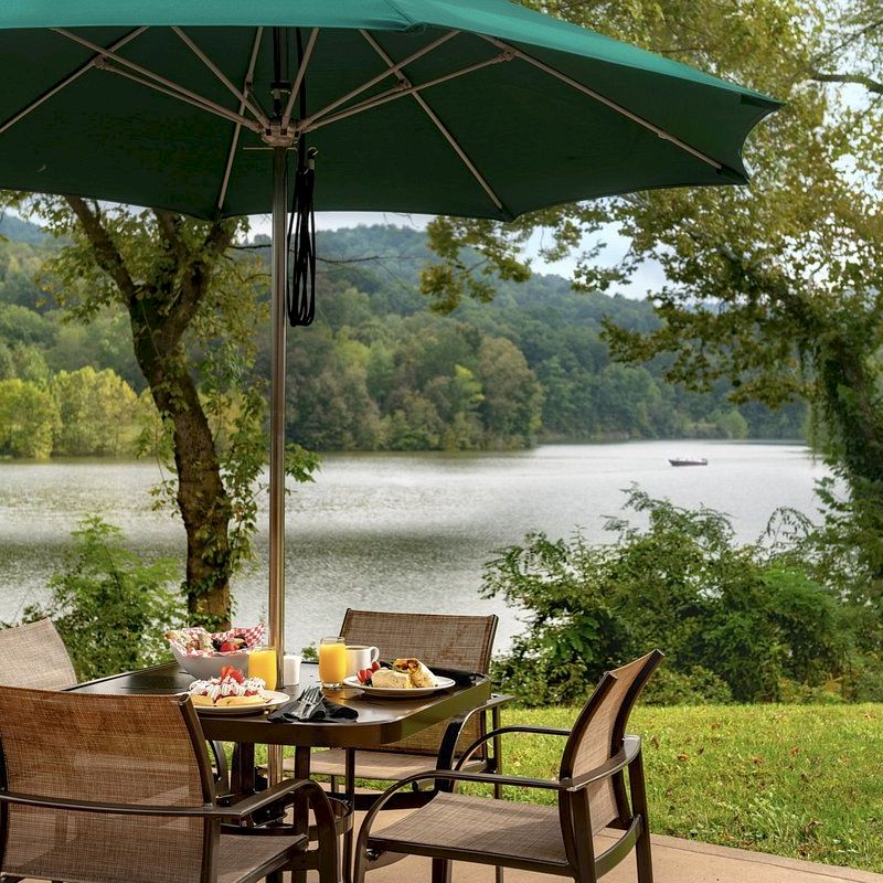 A patio set with a green umbrella, set for a meal, is overlooking a serene lake and wooded landscape under a partly cloudy sky.