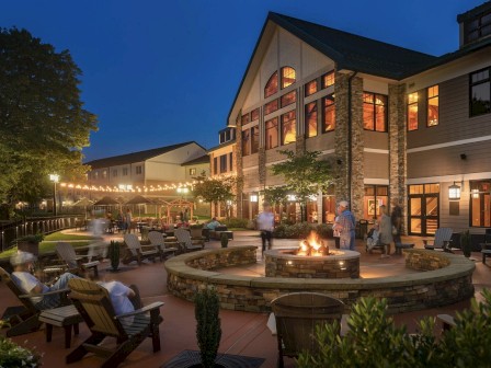 An outdoor patio with lounge seating and a central firepit, surrounded by a large building with many windows, all lit up at dusk.