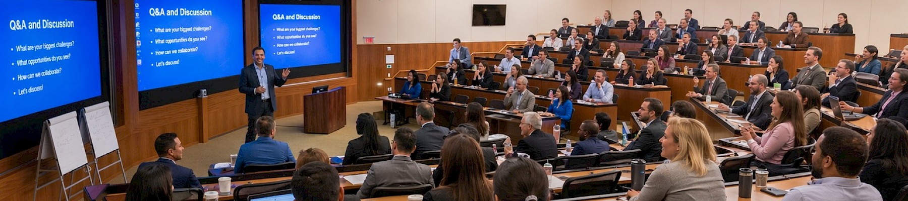 A large, professional conference or seminar with a speaker presenting to an attentive, diverse audience in a modern, well-lit lecture hall.