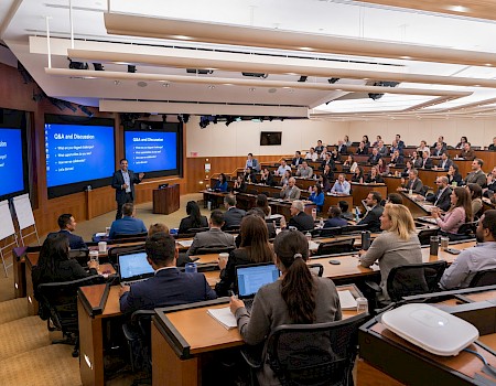 A large, professional conference or seminar with a speaker presenting to an attentive, diverse audience in a modern, well-lit lecture hall.