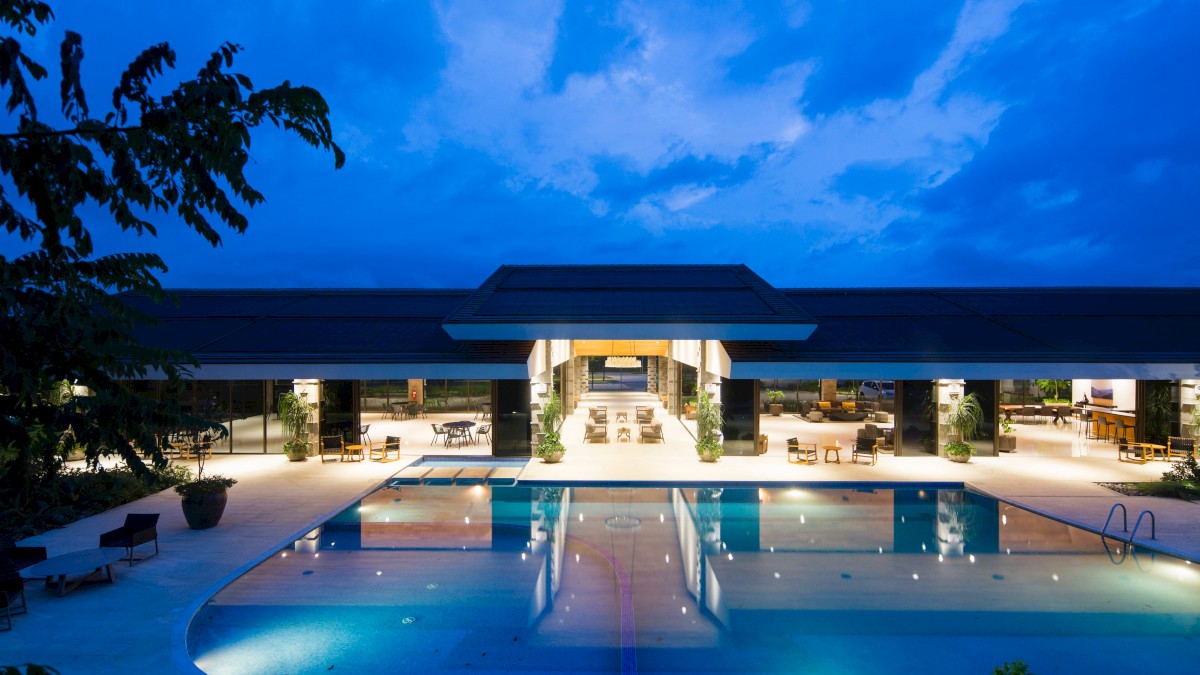 The image shows a beautifully lit outdoor swimming pool in front of a modern building with open-air seating, set against a twilight sky.