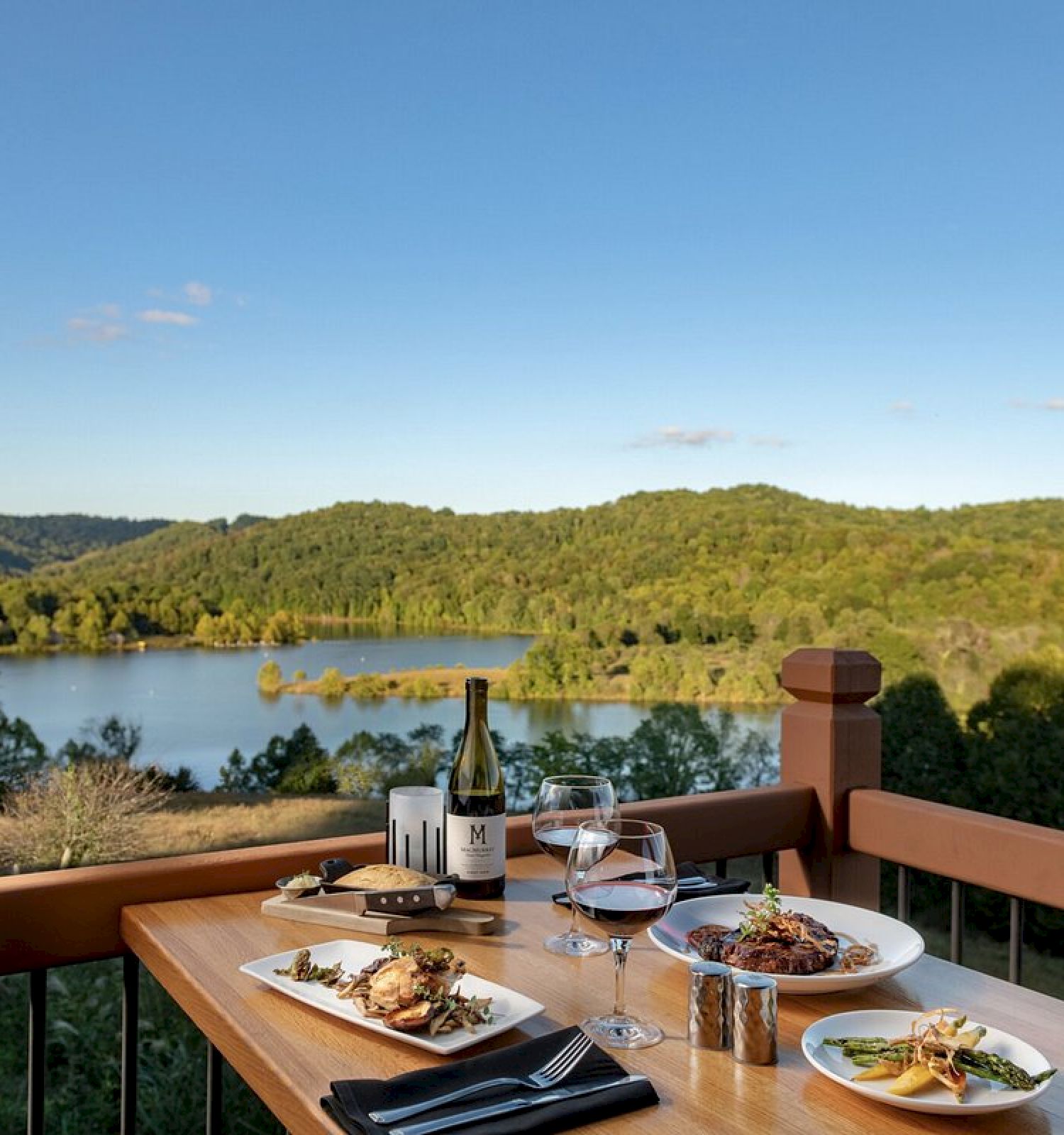 A table with a meal and wine overlooks a scenic lake and lush green hills on a clear day, with blue sky and a few scattered clouds in the background.