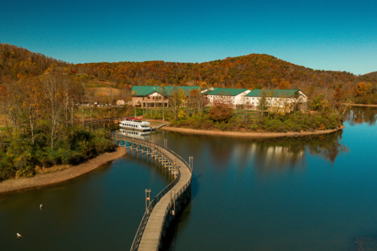 An aerial view of a serene lake with a footbridge, surrounded by autumn trees, leading to buildings with green roofs, against a backdrop of hills.