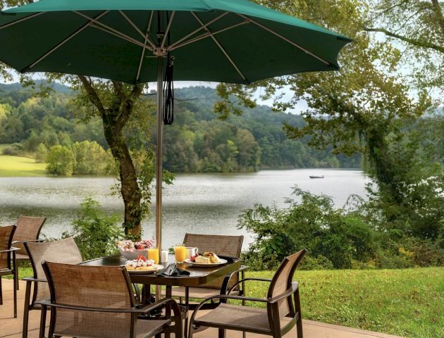 An outdoor patio set with a green umbrella, chairs around a table with breakfast items, overlooking a serene lake and forested hills.