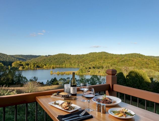 A wooden table on a balcony with a picturesque lake and forest view, set with plates of food, glasses of wine, and a bottle, under a clear blue sky.