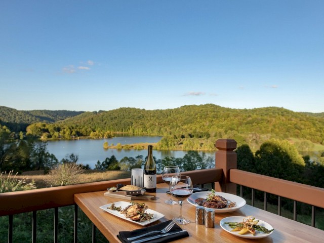 An outdoor dining setup with plates of food, wine, and glasses on a wooden table, overlooking a serene lake and surrounding green hills.