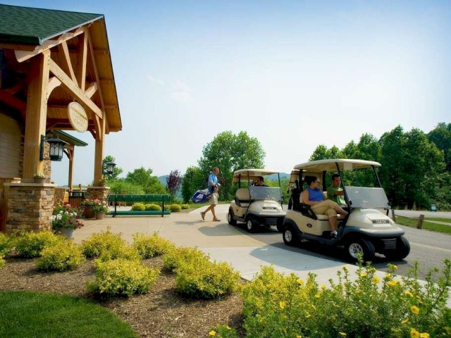Two people are seated in golf carts outside a wooden building, with another person walking nearby carrying a golf bag. The area is landscaped.