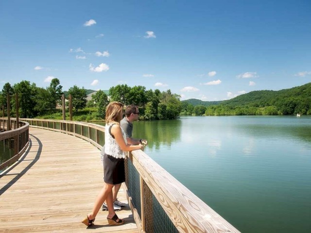 Two people are standing on a wooden boardwalk over a serene lake, surrounded by lush green trees and hills under a clear blue sky.