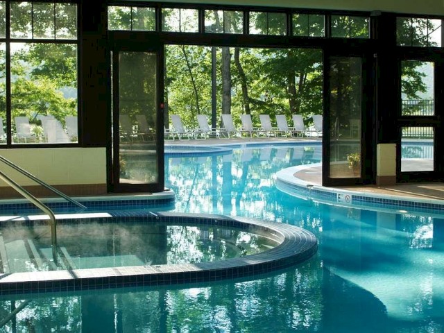 The image shows an indoor swimming pool area with an outdoor view, featuring clear water, lounge chairs, and large windows letting in natural light.