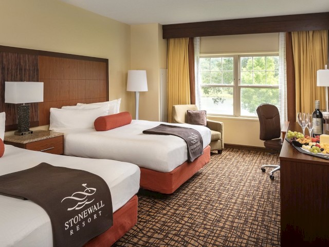 A hotel room with two double beds, a chair near the window, a desk with wine and a fruit plate, and lamps on nightstands and a floor lamp.