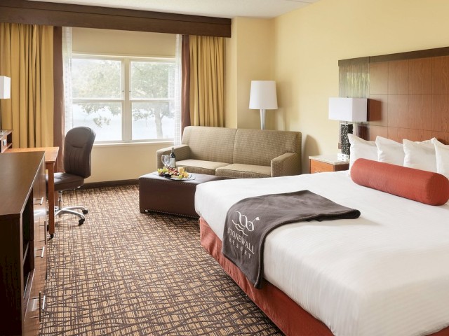 This is a modern hotel room featuring a large bed, a desk with a chair, a flat-screen TV, a sofa, and a window with curtains.