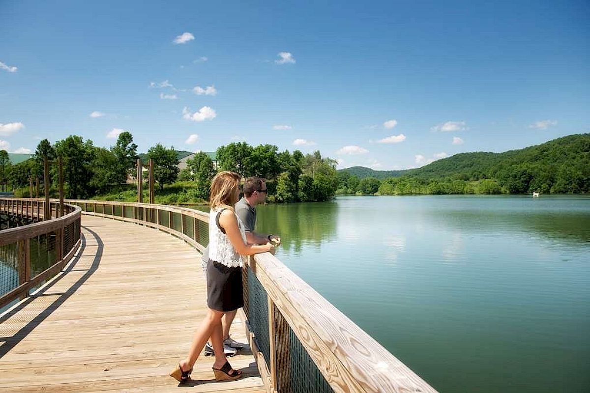 A couple stands on a wooden boardwalk overlooking a serene lake surrounded by lush green trees and hills under a clear blue sky and fluffy clouds.