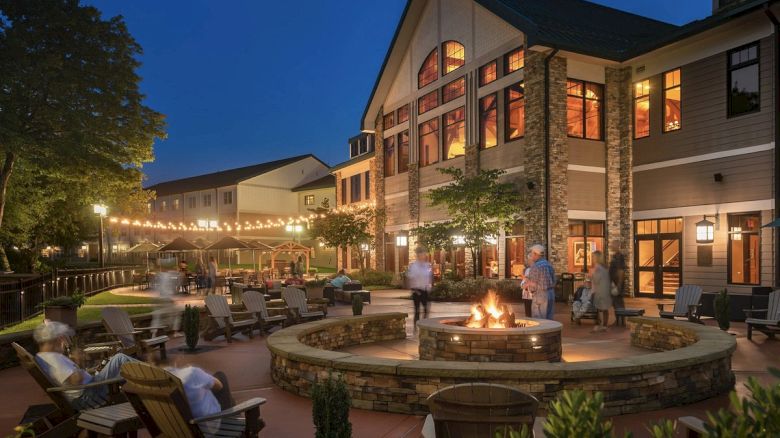 A nighttime scene shows people relaxing around an outdoor fire pit near a modern building, illuminated by string lights and surrounded by greenery.