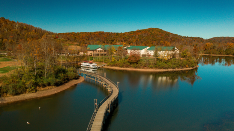 A serene lake scene with a curved wooden bridge leading to a docked boat, and buildings surrounded by autumn trees reflecting on the water under a clear sky.