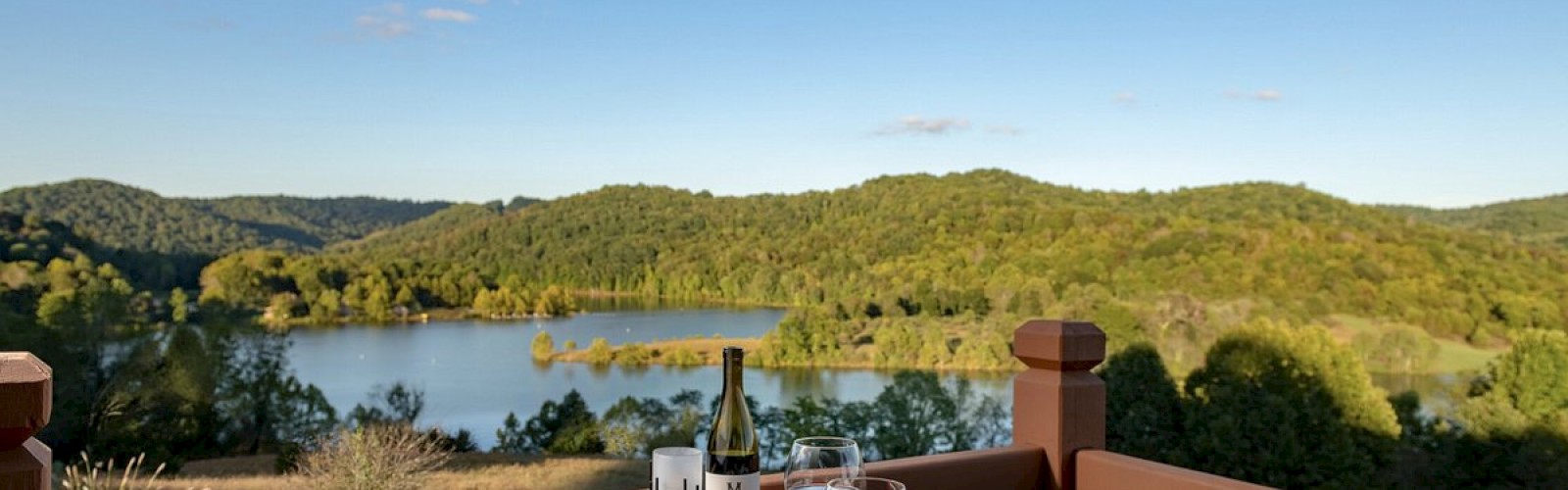 A table set with food, wine, and glasses on a balcony overlooking a scenic lake and hills under a clear blue sky.