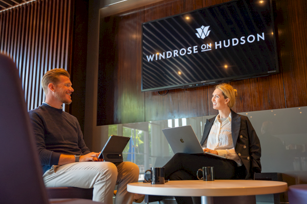 Two professionals sit and chat in a stylish office lounge, engaging in a friendly discussion with laptops.