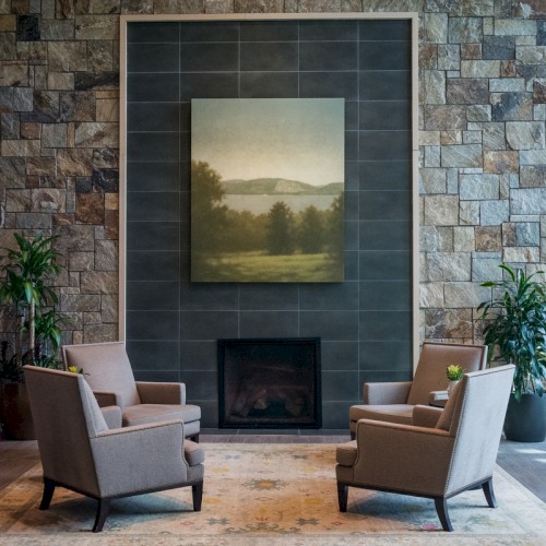 A cozy seating area with four beige armchairs around a fireplace, flanked by potted plants and a large landscape painting above.