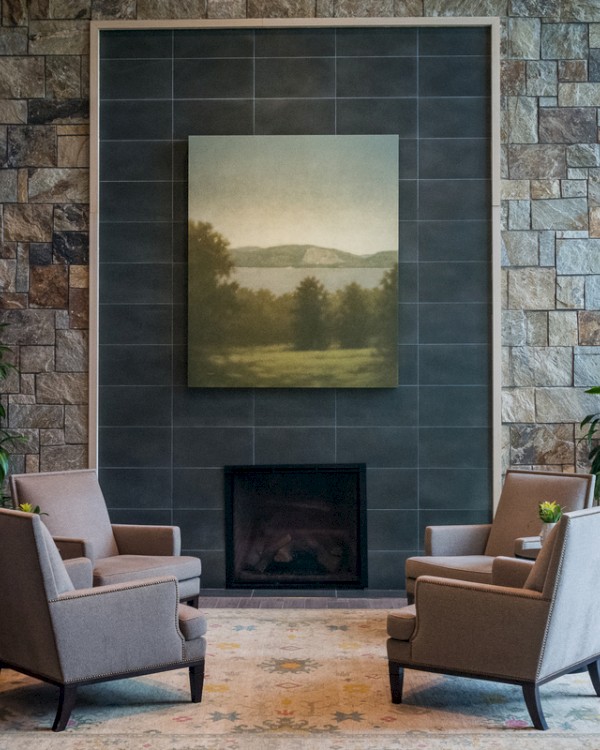 A cozy seating area with four beige armchairs around a fireplace, flanked by potted plants and a large landscape painting above.