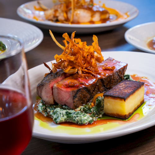 A beautifully plated steak with crispy onions, creamy spinach, a rectangular potato cake, and a glass of red wine.