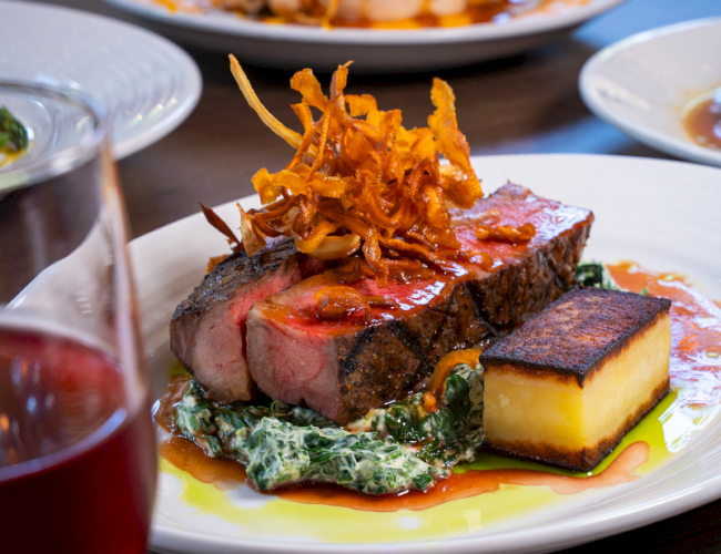 A beautifully plated steak with crispy onions, creamy spinach, a rectangular potato cake, and a glass of red wine.