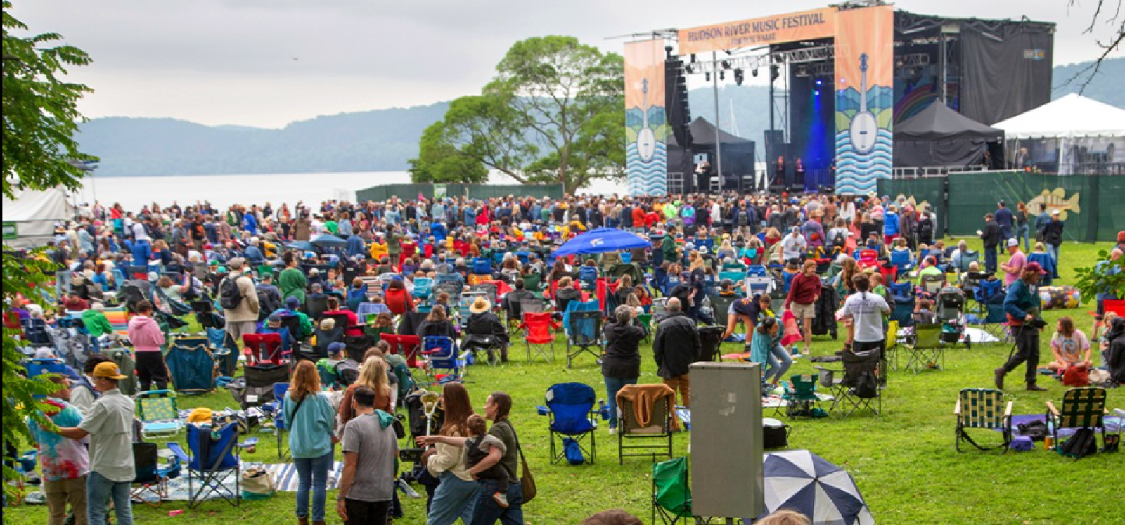 A large outdoor music festival by a lake with a seated crowd, a stage, and tents, where people enjoy the event on a lush green field.