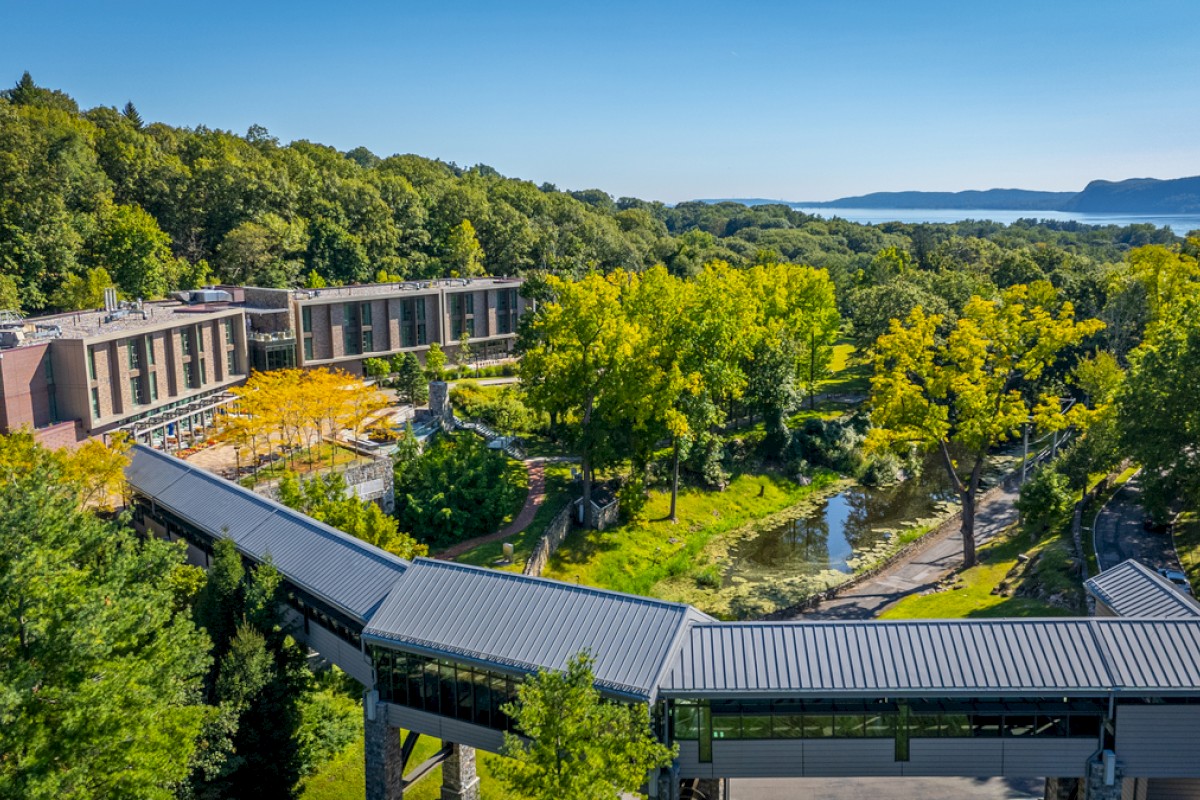 A scenic view of a modern building surrounded by lush trees, a pond, and distant hills in a peaceful natural setting.
