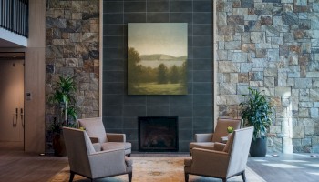 A cozy seating area with four beige armchairs around a fireplace, flanked by potted plants and a large landscape painting above.