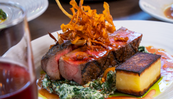 A beautifully plated steak with crispy onions, creamy spinach, a rectangular potato cake, and a glass of red wine.