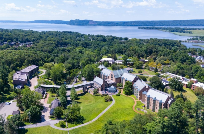 The image shows a scenic view of a university campus surrounded by trees, with a river and hills in the background on a clear day.