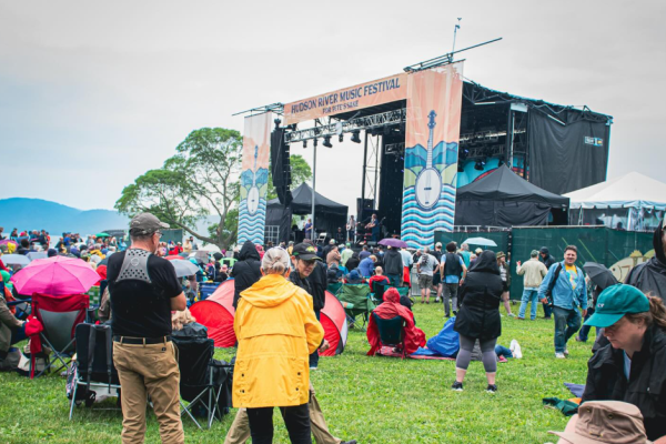 People enjoying a music festival outdoors with tents, umbrellas, and a large stage for the Hudson River Music Festival.