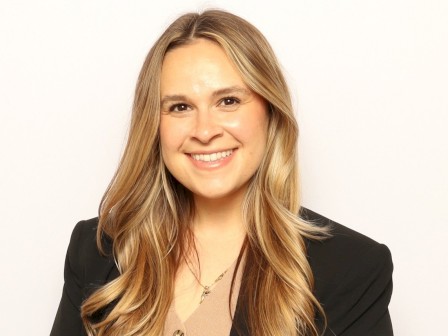 A smiling woman with long blonde hair, wearing a black blazer over a beige and black striped dress, posed in front of a plain white background.